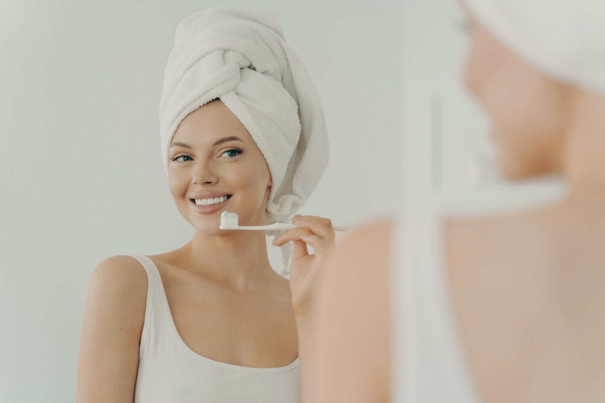 Beautiful young woman with healthy perfect smile brushing teeth and looking in mirror